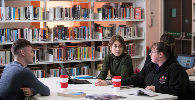 Three students working together at a desk in the library, with book shelves surrounding them.