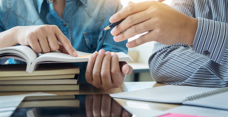 Two students are pointing at something in an open textbook, on top of a stack of closed textbooks.