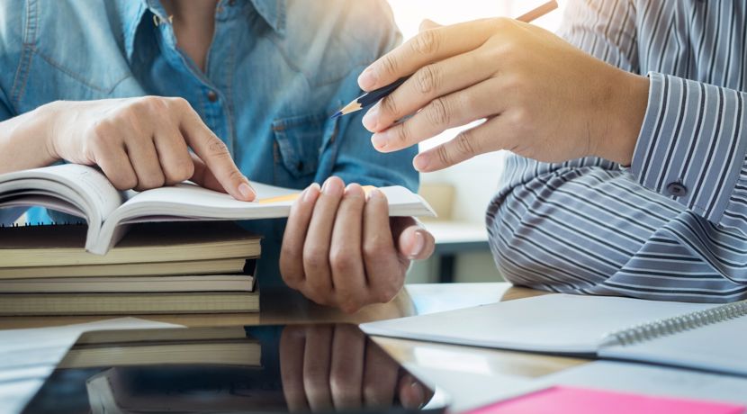 Two students are pointing at something in an open textbook, on top of a stack of closed textbooks.