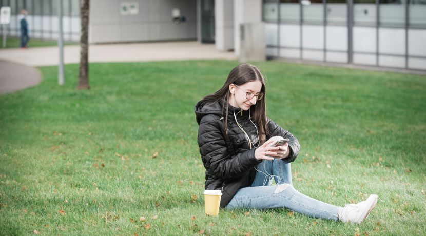 A young student is sitting on the grass looking at their phone at Granton campus. 